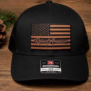 May include: Black trucker hat with a brown leather patch featuring an American flag design and the words "Raised American." The hat is adjustable and has a mesh back. The Richardson brand logo is visible.
