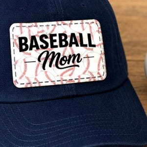 May include: A navy blue baseball cap with a rectangular patch that reads "BASEBALL Mom" in black and red lettering. The patch has a baseball pattern background. A baseball is visible on the right.
