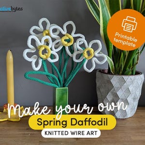 Op de afbeelding: Een handgemaakt draadkunst narcissenboeket. De bloemen hebben witte bloemblaadjes met gele centra en groene stelen, tentoongesteld in een groene vaas. De afbeelding bevat tekst die zegt "Make your own Spring Daffodil KNITTED WIRE ART". Een gele kaars staat links.