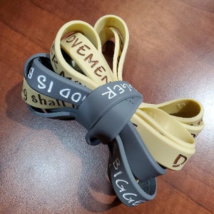 May include: Two silicone wristbands with the text "BIGGER" and "MOVEMENT IS DO" printed on them. The wristbands are in gray and beige.