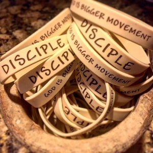 May include: A collection of off-white rubber wristbands with the word "DISCIPLE" and the phrase "GOD IS BIGGER MOVEMENT" printed in black. The bands are piled in a wooden bowl, creating a textured display.