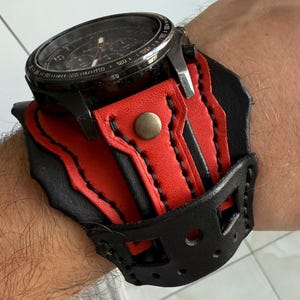 May include: A black and red leather watch cuff with a silver watch. The cuff has a red interior and black exterior with decorative cutouts and a snap closure. The watch has a black face and silver bezel.