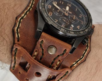 May include: A black wristwatch with a brown leather cuff. The watch face is dark with gold-colored hands and numerals. The cuff is made of distressed brown leather with decorative stitching and a metal button clasp.