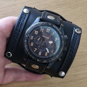 May include: A black Slazenger chronograph watch featuring a round face with copper-colored numerals and hands, set on a wide black leather cuff strap. The cuff is detailed with braided leather edges and four silver-toned metal rivets.