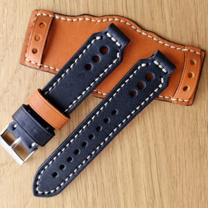 May include: Black and orange leather watch strap with white stitching and holes. The strap is shown with a spare piece of leather.