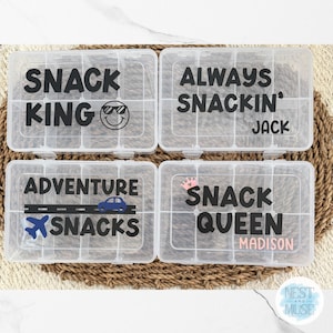 May include: Four clear plastic snack boxes with black text and graphics. The boxes read "SNACK KING", "ALWAYS SNACKIN' JACK", "ADVENTURE SNACKS", and "SNACK QUEEN MADISON". The boxes are divided into compartments.
