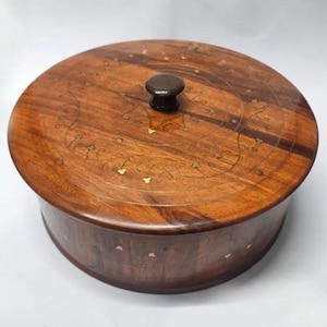 May include: A circular wooden box with a lid. The box is crafted from dark brown wood, featuring a lighter wood inlay design on the lid. A small, dark brown knob adorns the lid. The box is likely for storage.