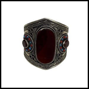 May include: Silver cuff bracelet with a central oval, deep red stone. The bracelet has decorative patterns and is accented with red and blue circular embellishments. A striking statement piece.