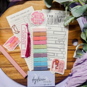 May include: A collection of stationery items for book lovers, including stickers, bookmarks, and colorful sticky notes. The items are arranged on a wooden board, with a pink pen and decorative greenery. The stickers have text that reads "Open to Book Recs" and "Leaving Reality."