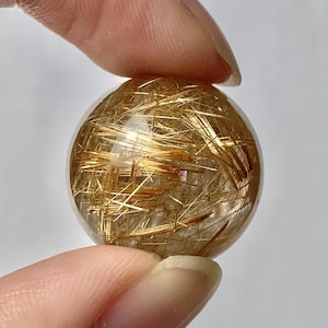 May include: A clear, spherical crystal ball with golden, needle-like inclusions. The crystal is held between two fingers, showcasing its transparency and the intricate patterns within. The golden needles create a striking visual effect.