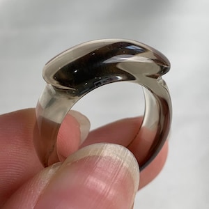 May include: A smooth, translucent smoky quartz ring featuring a polished, rounded band and a slightly elevated, elongated oval top section. The gemstone exhibits a deep, semi-transparent brown hue with natural variations in clarity and light reflection.