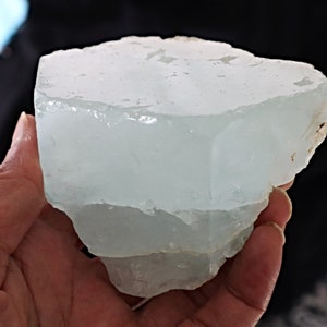 May include: A large, translucent, light blue aquamarine crystal with a rough, natural texture. The crystal is shaped like a block with several layers.