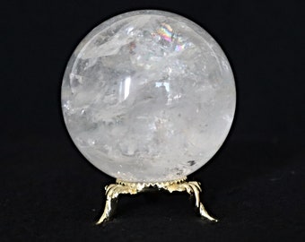 May include: A clear quartz crystal ball, approximately 4 inches in diameter, sits on a decorative gold-colored metal stand. The crystal sphere has internal fractures and displays rainbow refractions. The background is solid black.