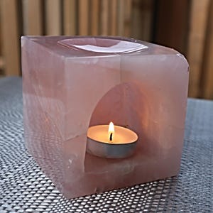 May include: A pink rose quartz candle holder with a small white tea light candle inside. The candle holder has a square shape with a cutout in the center for the candle.