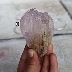 May include: A large, translucent, pink and white crystal with striations. The crystal has a rough, natural texture and is held in a hand.