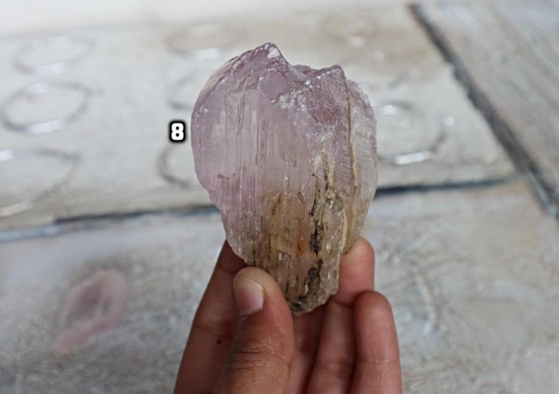 May include: A large, translucent, pink and white crystal with striations. The crystal has a rough, natural texture and is held in a hand.