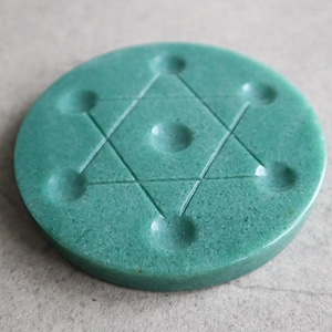 May include: A circular, teal-coloured stone featuring a carved Star of David design. The stone has a textured surface and six small, circular indentations around the star. Likely used for spiritual or decorative purposes.