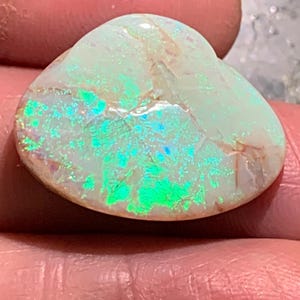 Puede incluir: Una piedra preciosa de ópalo pulido con una base de color blanco cremoso. El ópalo muestra destellos vibrantes de iridiscencia verde y azul en su superficie. La piedra se sostiene en una mano, mostrando su forma natural y su juego de colores único.