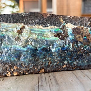 May include: A roughly rectangular stone with a dark, textured exterior. The interior displays vibrant blues, greens, and iridescent colours, suggesting an opal or similar gemstone. The stone rests on a wooden surface.