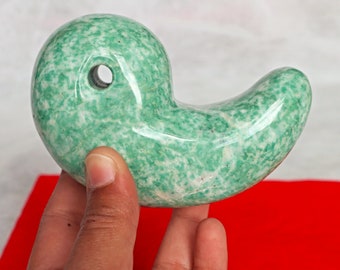 Puede incluir: Una talla de piedra de jade verde en forma de pájaro estilizado con un agujero en el centro. La piedra tiene un patrón moteado verde y blanco.
