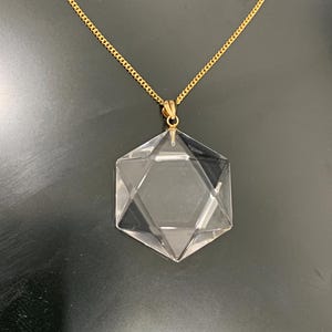May include: A clear crystal pendant in the shape of a Star of David hangs from a gold-colored chain. The pendant is faceted, reflecting light, and the chain is a delicate curb style. The background is a dark, solid color.