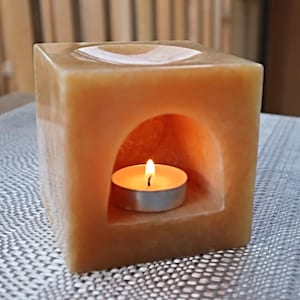 May include: A square, light orange, stone diffuser with a small, round, white candle inside. The candle is lit and the stone has a small opening in the front.