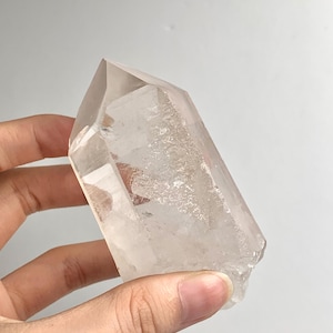 May include: A clear, hexagonal quartz crystal point with a pointed termination. The crystal features a smooth, polished surface on one side and a textured, cloudy interior with natural inclusions and facets visible throughout the structure.