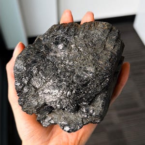 Puede incluir: Un gran trozo de carbón de forma irregular, sostenido en una mano. El carbón es de color negro intenso con una superficie brillante y reflectante. La textura parece áspera y desigual, con grietas y fisuras visibles. El fondo está borroso.