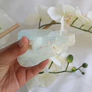 May include: A large, light blue aquamarine crystal with a rough, natural texture. The crystal is translucent and has a slight green hue. It is being held in a hand.