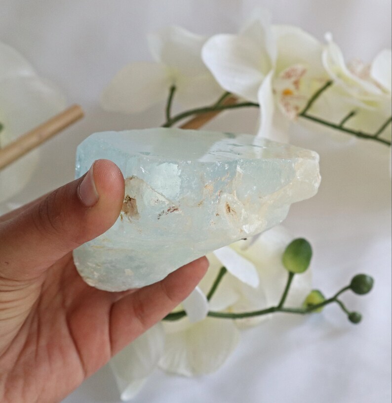 May include: A large, light blue aquamarine crystal with a rough, natural texture. The crystal is translucent and has a slight green hue. It is being held in a hand.