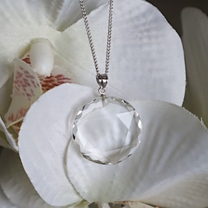 May include: A clear, faceted crystal pendant necklace with a silver chain. The round pendant is suspended from a delicate chain. The necklace is displayed against a backdrop of white flower petals.