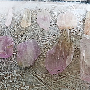 May include: A collection of pink and clear crystals, some with a slight purple hue, arranged on a white textured surface. The crystals are various shapes and sizes, with some being long and thin, while others are more rounded.