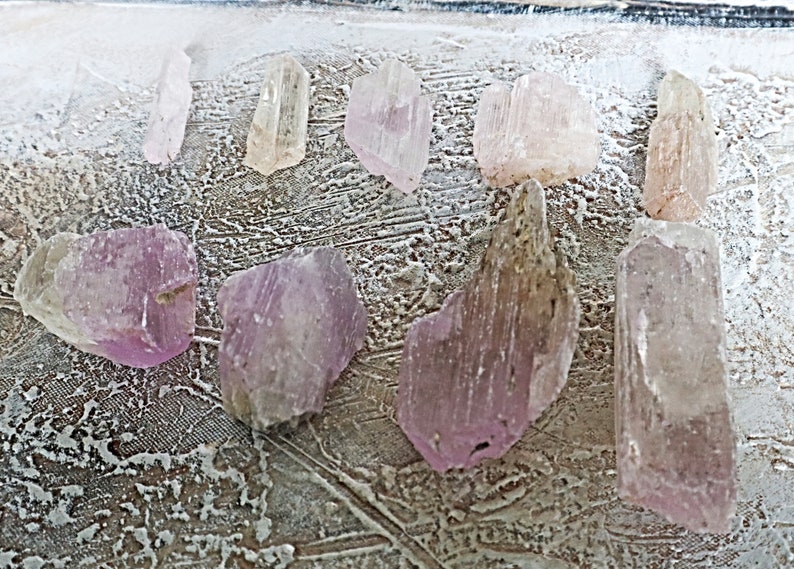May include: A collection of pink and clear crystals, some with a slight purple hue, arranged on a white textured surface. The crystals are various shapes and sizes, with some being long and thin, while others are more rounded.