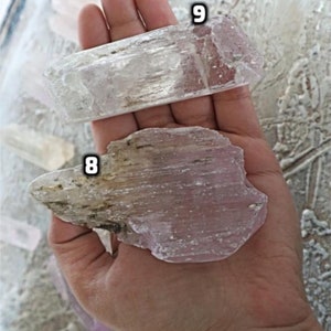 May include: Two pieces of selenite crystal, one clear and one with a pink hue. The crystals are both flat and have a striated texture. The crystals are labeled with the numbers 8 and 9.