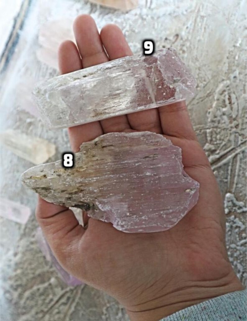 May include: Two pieces of selenite crystal, one clear and one with a pink hue. The crystals are both flat and have a striated texture. The crystals are labeled with the numbers 8 and 9.