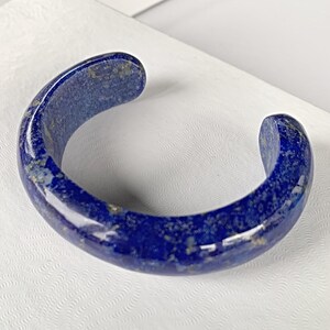 May include: A polished, open-ended cuff bracelet in a deep blue hue, crafted from a stone with gold flecks. The bracelet has a smooth, curved design, showcasing the natural beauty of the material. This piece of jewelry is a statement accessory.