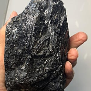 May include: A large, irregularly shaped, black mineral specimen with a glossy, reflective surface. The rough texture and dark color suggest a natural, unpolished stone, possibly a type of coal or obsidian. Held in a hand.
