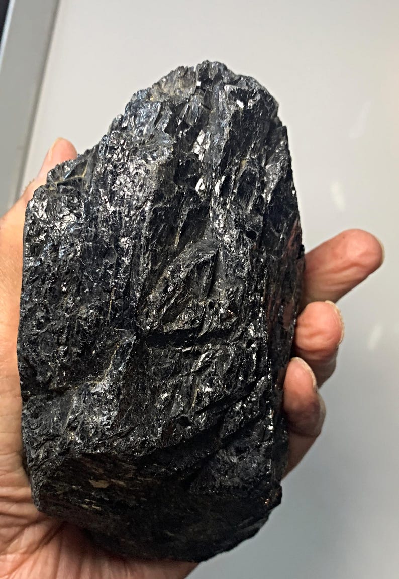 May include: A large, irregularly shaped, black mineral specimen with a glossy, reflective surface. The rough texture and dark color suggest a natural, unpolished stone, possibly a type of coal or obsidian. Held in a hand.