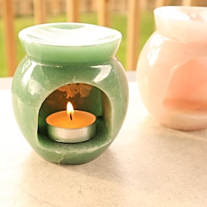 Può includere: Due scaldatori di olio decorativi, uno verde e uno rosa. Lo scaldatore verde ha una candela tealight accesa all'interno. Entrambi hanno un'apertura rotonda e un coperchio. Sono su una superficie bianca.