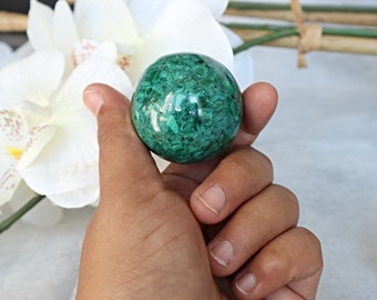 May include: A green malachite sphere, polished and smooth, with intricate patterns and swirls. The sphere is held in a hand, showcasing its size and detail.