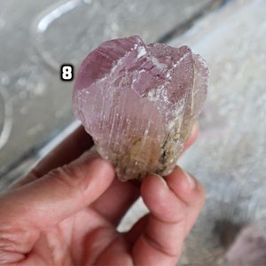 May include: A large, pink, and clear crystal with a rough, natural texture. The crystal has a light pink hue and is translucent. The crystal is labeled with the number "8".