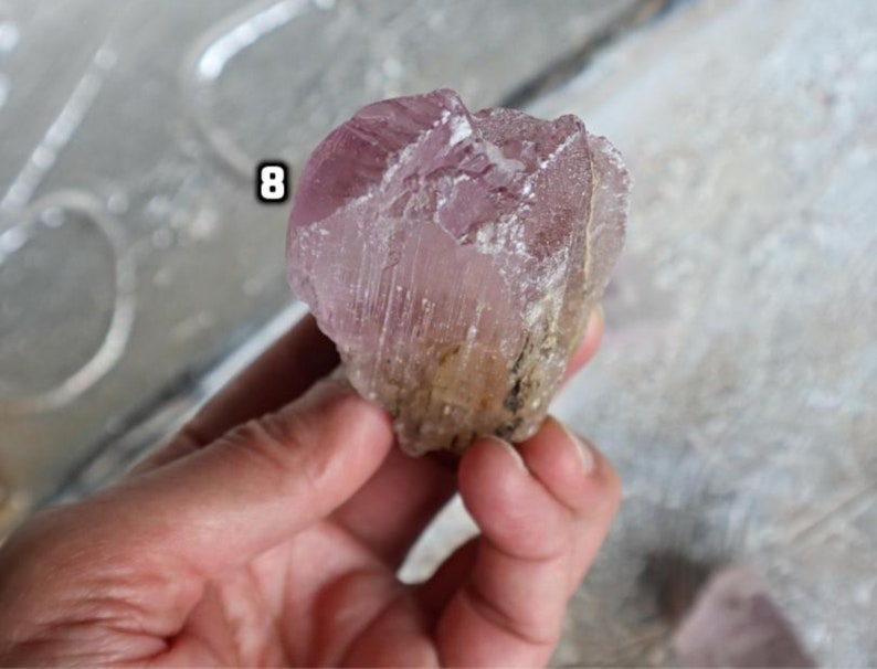 May include: A large, pink, and clear crystal with a rough, natural texture. The crystal has a light pink hue and is translucent. The crystal is labeled with the number "8".