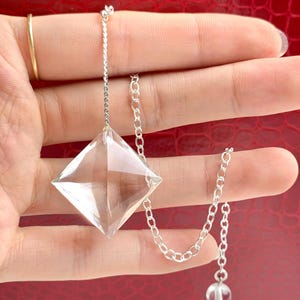 Puede incluir: Un colgante de cristal transparente en forma de diamante suspendido de una cadena de metal plateado. La cadena presenta una pequeña cuenta de cristal esférica y transparente en el extremo. El colgante tiene una superficie facetada.