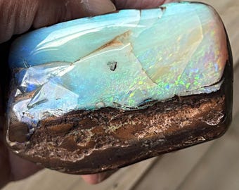 May include: A polished opal stone with a vibrant display of colours. The top surface showcases a gradient of blues and greens, with flashes of iridescent hues. The bottom portion is a rough, earthy brown, creating a striking contrast.