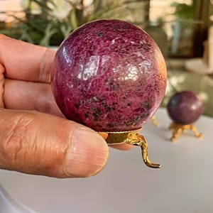 Puede incluir: Una esfera de piedra preciosa magenta oscuro pulida y redonda, con motas oscuras, sostenida en una mano. La esfera se asienta sobre un pequeño soporte dorado ornamentado. Otra esfera es visible en el fondo.