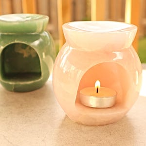 May include: Two ceramic oil warmers. One is a pale green colour, the other is a soft pink. The pink warmer has a lit tea light candle inside. Both have a round shape with a large opening in the front.