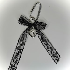 Peut inclure: Un mousqueton argent&eacute; avec un n&oelig;ud en dentelle noire et un pendentif en forme de c&oelig;ur. Le ruban de dentelle s'&eacute;tend vers le bas. Le design est &eacute;l&eacute;gant et gothique, adapt&eacute; aux porte-cl&eacute;s ou aux accessoires de sac.