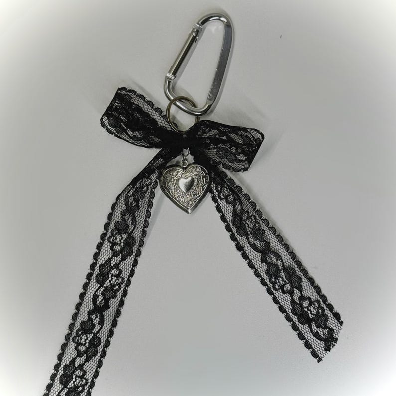 Peut inclure: Un mousqueton argent&eacute; avec un n&oelig;ud en dentelle noire et un pendentif en forme de c&oelig;ur. Le ruban de dentelle s'&eacute;tend vers le bas. Le design est &eacute;l&eacute;gant et gothique, adapt&eacute; aux porte-cl&eacute;s ou aux accessoires de sac.