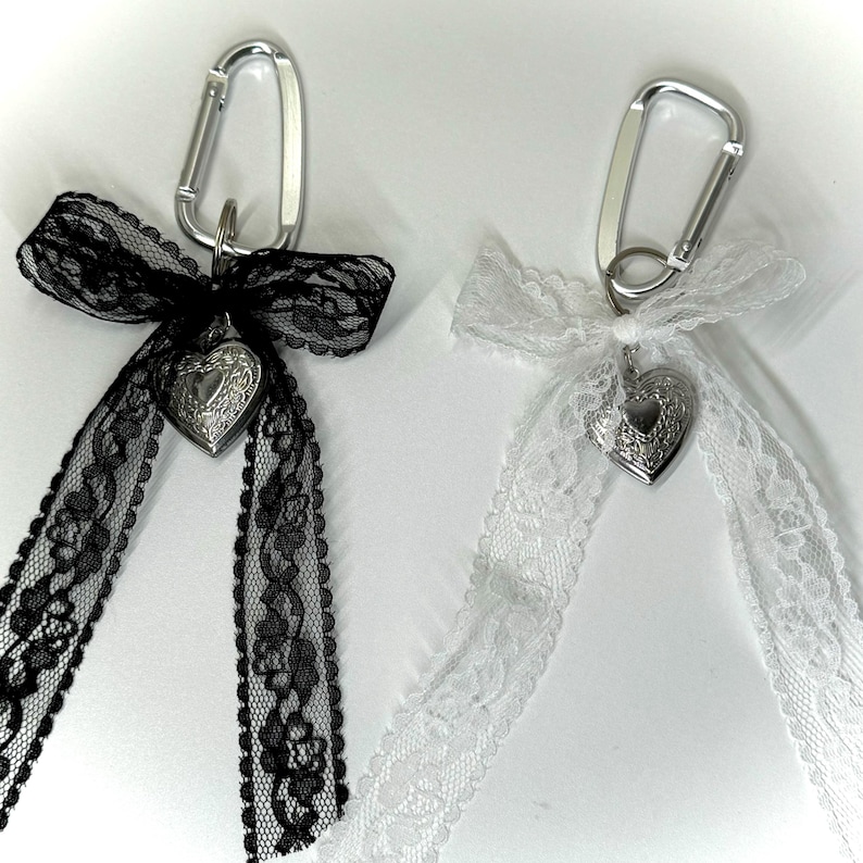 Peut inclure: Deux porte-cl&eacute;s avec mousquetons argent&eacute;s. L'un a un ruban de dentelle noire et un m&eacute;daillon en forme de c&oelig;ur. L'autre a un ruban de dentelle blanche et un m&eacute;daillon en forme de c&oelig;ur. Sur fond blanc.