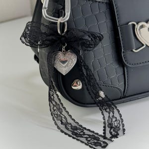 Peut inclure: Sac &agrave; main noir en similicuir avec une texture crocodile. Le sac est orn&eacute; d'un fermoir argent&eacute; en forme de c&oelig;ur et d'un n&oelig;ud d&eacute;coratif en dentelle noire avec un pendentif en forme de c&oelig;ur. Un mousqueton argent&eacute; est attach&eacute; en haut.